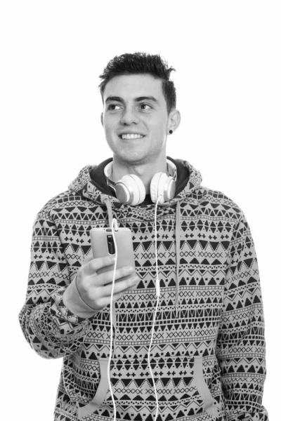 Jeune homme heureux réfléchi souriant et tenant le téléphone portable tout en portant des écouteurs autour du cou — Photo
