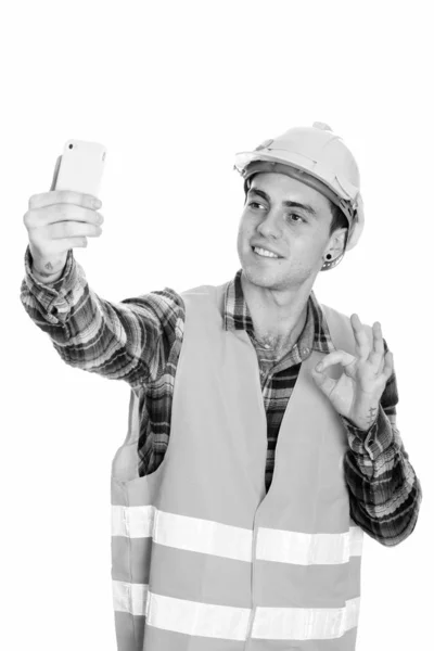 Jovem feliz trabalhador da construção tomando selfie e dando sinal ok — Fotografia de Stock