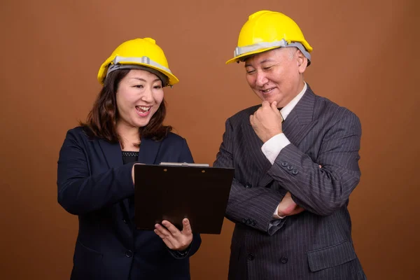 成熟した日本人実業家と成熟した日本人実業家が茶色の背景を背景に一緒にハードハットをかぶってスタジオショット — ストック写真