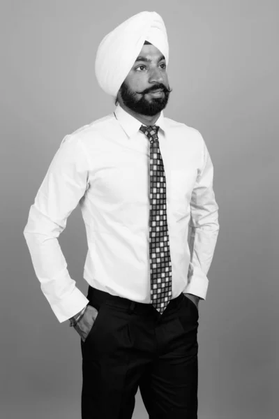 Studio shot of young bearded Indian Sikh businessman with turban against gray background in black and white