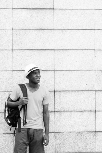 Feliz joven turista africano pensando y sosteniendo la mochila contra la pared de bloque de hormigón —  Fotos de Stock