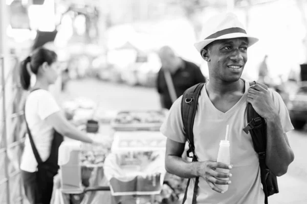 Young happy African tourist man thinking while holding bottle of orange juice in the streets — Stock Photo, Image