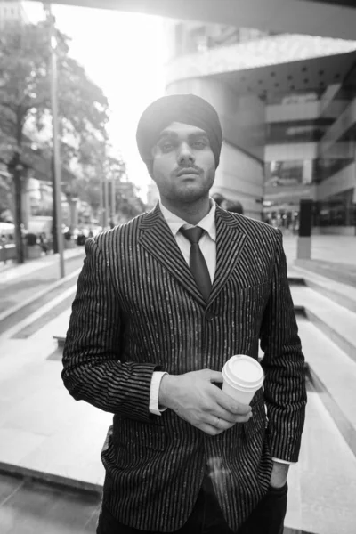 Portrait of young handsome Indian Sikh businessman wearing turban while exploring the city in black and white