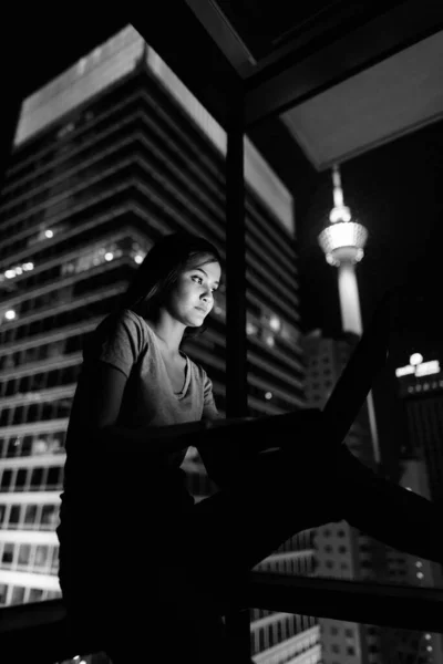 年轻美丽的亚洲女子的画像 她穿着黑白相间的夜景 靠着玻璃窗 用笔记本电脑看城市 — 图库照片