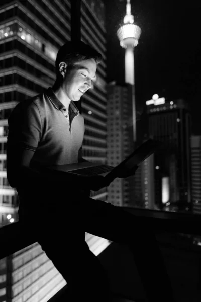 年轻英俊的人的肖像使用笔记本电脑对玻璃窗与城市的看法在晚上在黑色和白色 — 图库照片