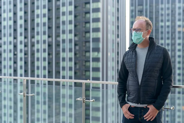 Portrait of mature Scandinavian man wearing mask for protection from corona virus outbreak and pollution against view of the city