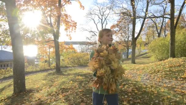 Ευτυχισμένο νεαρό όμορφο αγόρι που πετάει φθινοπωρινά φύλλα στο πάρκο — Αρχείο Βίντεο