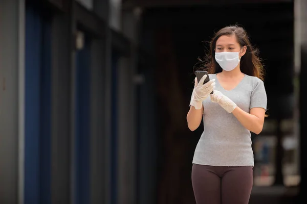 Fiatal ázsiai nő használ telefon maszk és kesztyű szabadban — Stock Fotó