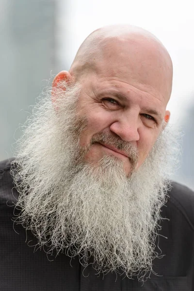 Portrait of mature bald hipster man with long beard against view of the city outdoors