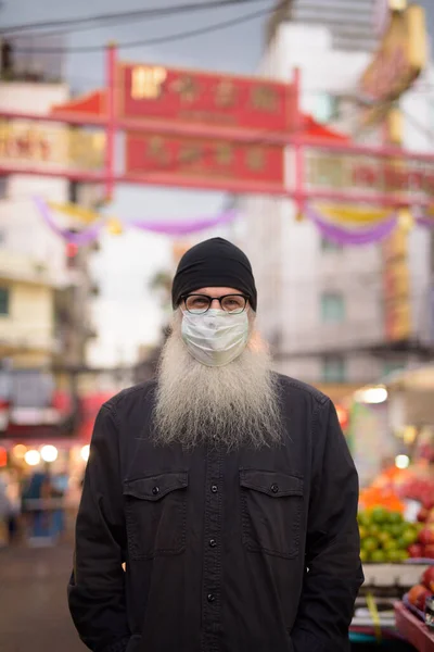 Portrait of mature bearded tourist man with mask for protection from corona virus outbreak in Chinatown at Bangkok city