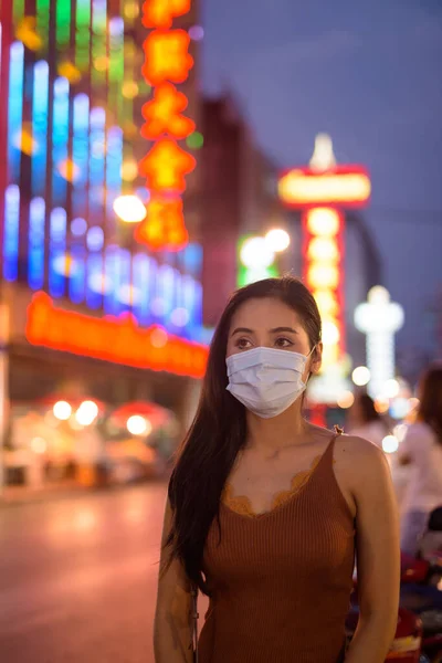 Portrait of young beautiful Asian tourist woman with mask for protection from corona virus outbreak in Chinatown at night