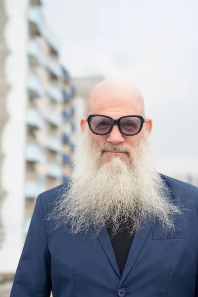 Portrait of mature handsome bald hipster businessman with long beard against view of the city