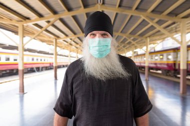 Tren istasyonundaki Corona virüsü salgınından korunmak için maskeli yetişkin sakallı hippi turist portresi.