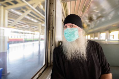 Korona virüsü salgınından korunmak için maske takan olgun sakallı turist portresi tren istasyonunda.