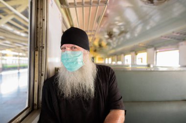 Korona virüsü salgınından korunmak için maske takan olgun sakallı turist portresi tren istasyonunda.