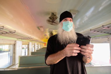Korona virüsü salgınından korunmak için maske takan olgun sakallı turist portresi tren istasyonunda.