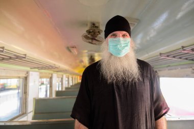 Korona virüsü salgınından korunmak için maske takan olgun sakallı turist portresi tren istasyonunda.