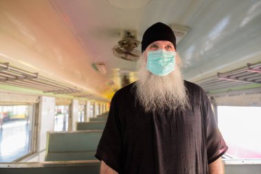 Korona virüsü salgınından korunmak için maske takan olgun sakallı turist portresi tren istasyonunda.