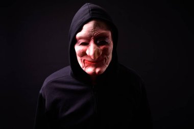Studio shot of young Asian man with hoodie and horror mask against black background