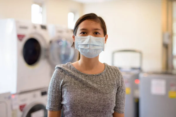 Portrait of young Filipino woman with mask for protection from corona virus outbreak at the laundromat indoors