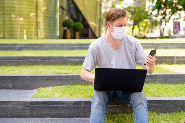 Portrait of young Scandinavian man with mask for protection from corona virus outbreak outside modern building with nature