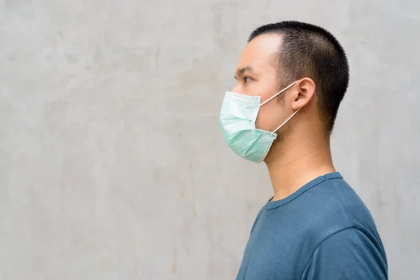 Portrait of young Asian man with mask for protection from corona virus outbreak against concrete wall outdoors