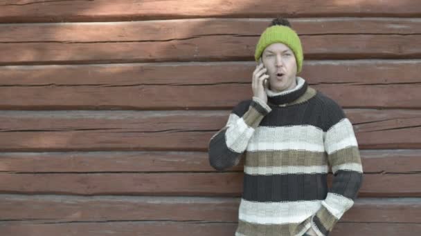 Szczęśliwy młody człowiek rozmawiający przez telefon pod drewnianą ścianą jesienią — Wideo stockowe