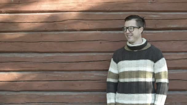 Vrolijke jongeman met een bril glimlachend tegen een houten muur in de herfst — Stockvideo