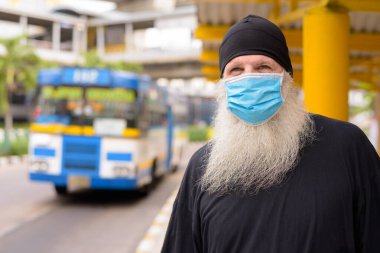 Korona virüsü salgınından ve otobüs durağındaki kirlilikten korunmak için maske takan olgun sakallı bir hippi.