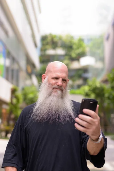 Mature handsome bald bearded man using phone with nature in the city