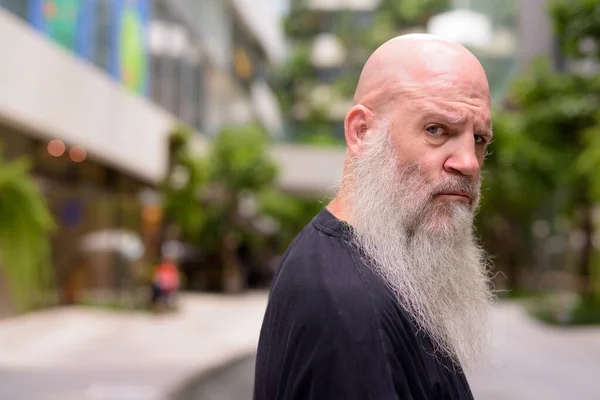 Face of mature bald bearded man looking serious at camera with nature in the city — Stock Photo, Image
