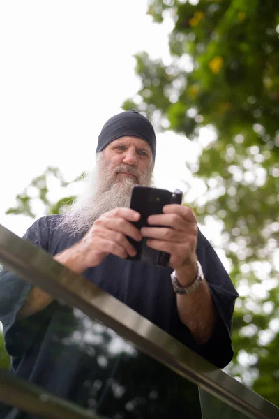 Homme hipster barbu mature regardant sérieusement tout en utilisant le téléphone à l'extérieur de la gare du métro — Photo