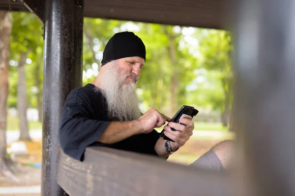 Volwassen knap bebaarde hipster man met behulp van telefoon terwijl zitten op houten bank buiten — Stockfoto