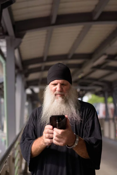 Rijpere knappe bebaarde hipster man met behulp van telefoon op footbridge in de stad — Stockfoto