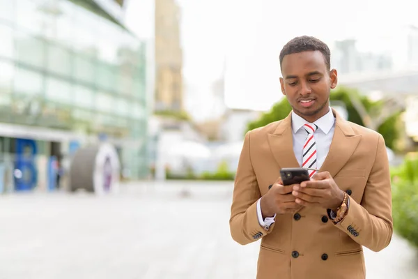 Happy young handsome African businessman using phone in the city outdoors — Stock Photo, Image