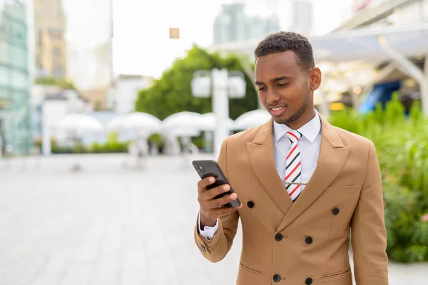 Happy young handsome African businessman using phone in the city outdoors — Stock Photo, Image