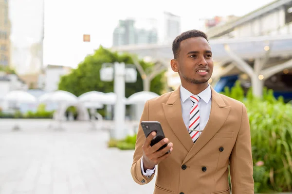 Feliz joven guapo empresario africano pensando mientras usa el teléfono en la ciudad — Foto de Stock