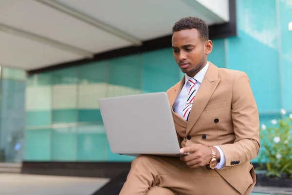 Joven hombre de negocios africano guapo usando el ordenador portátil mientras está sentado fuera del edificio — Foto de Stock