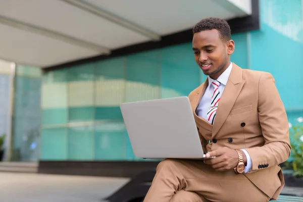 Feliz joven guapo empresario africano usando el ordenador portátil mientras está sentado en la ciudad al aire libre — Foto de Stock