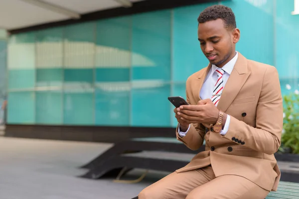 Jovem empresário africano bonito usando telefone sentado fora do prédio — Fotografia de Stock