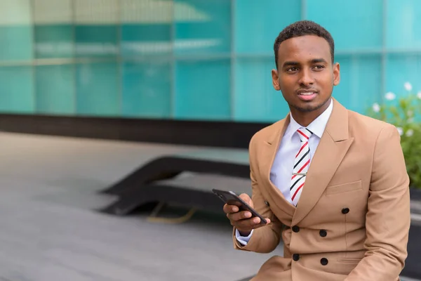 Feliz joven empresario africano con pensamiento telefónico y sentado en la ciudad al aire libre — Foto de Stock
