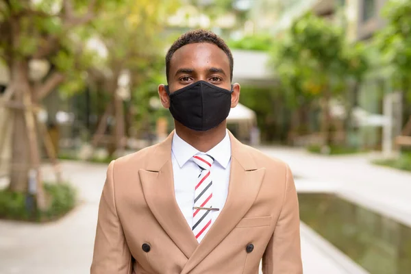 Молодой африканский бизнесмен в маске для защиты от вспышки коронного вируса в городе с природой — стоковое фото