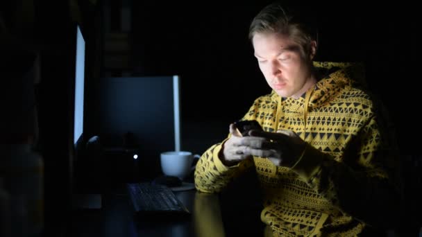 Joven usando el teléfono y tomando café mientras trabaja horas extras en casa en la oscuridad — Vídeos de Stock