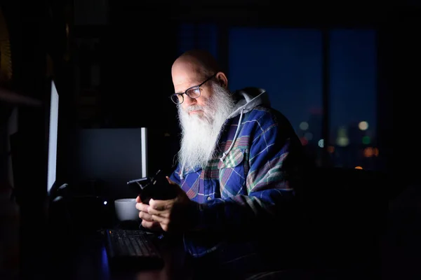 Portrait of mature handsome bald hipster man with long beard working overtime at home late at night in the dark