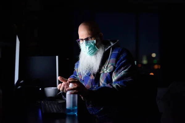 Mature bald bearded man with mask for protection from corona virus outbreak working from home during quarantine late at night