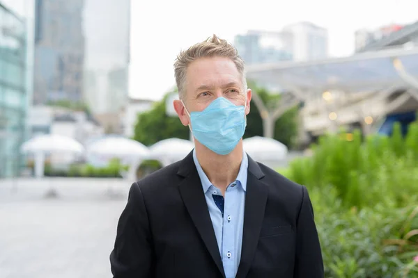 Portrait of mature businessman with mask for protection from corona virus outbreak against view of the city outdoors