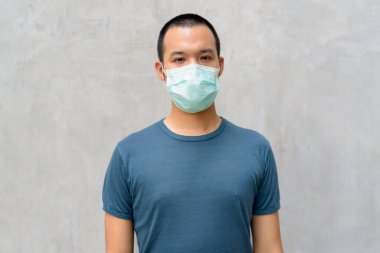 Portrait of young Asian man with mask for protection from corona virus outbreak against concrete wall outdoors
