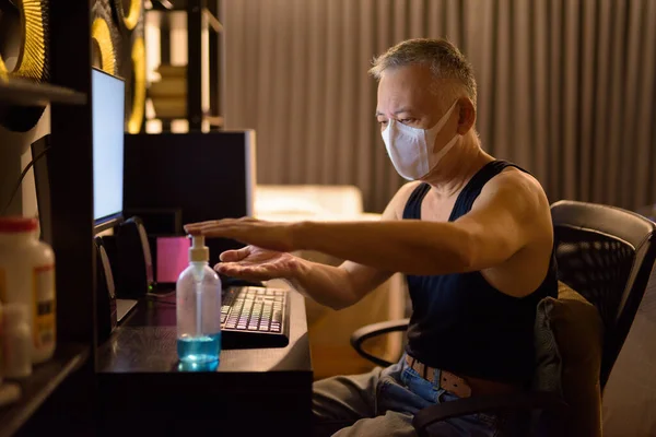 Portrait of mature Japanese man with mask for protection from corona virus outbreak working overtime at home late at night