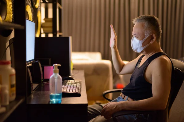 Portrait of mature Japanese man with mask for protection from corona virus outbreak working overtime at home late at night