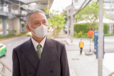 Portrait of mature Japanese businessman with mask for protection from corona virus outbreak in the city outdoors